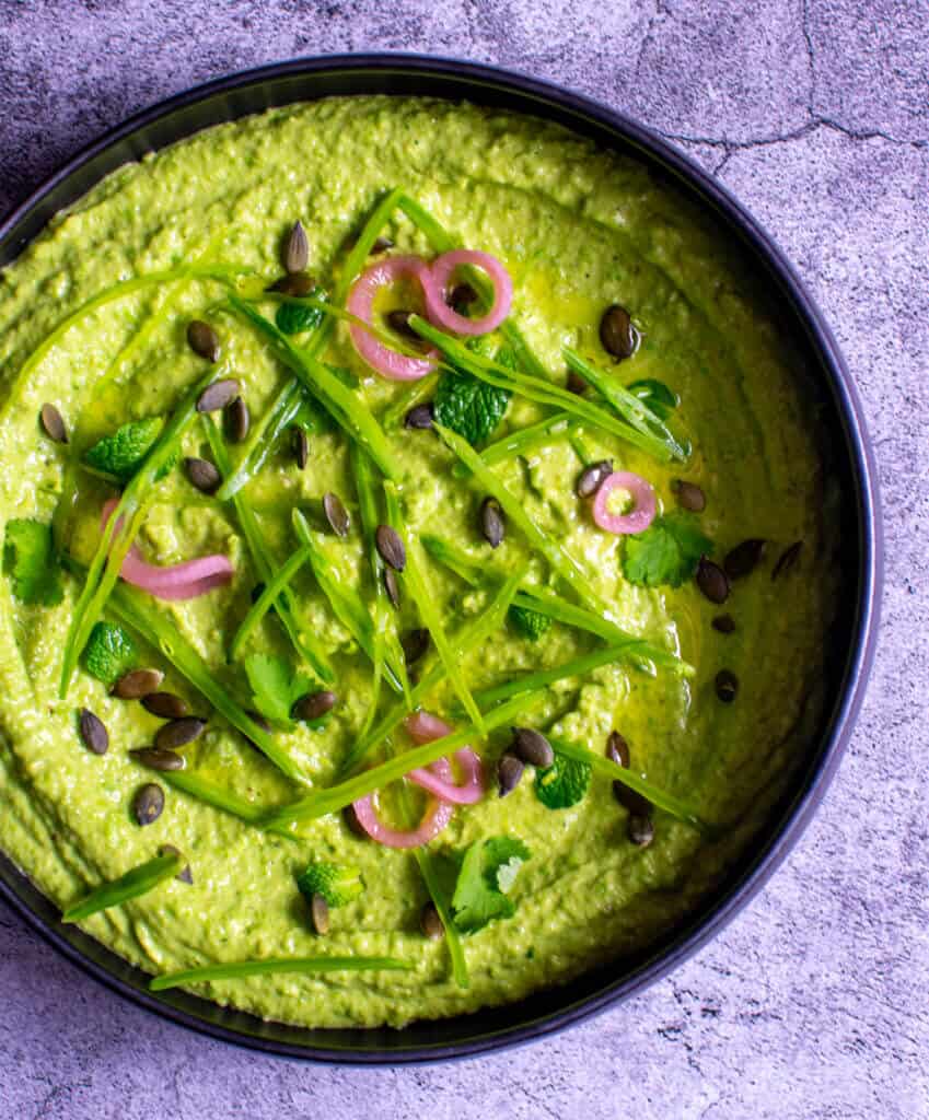 black bowl with avocado & pea hummus topped with pickled onions, pepitas, herbs & snowpeas