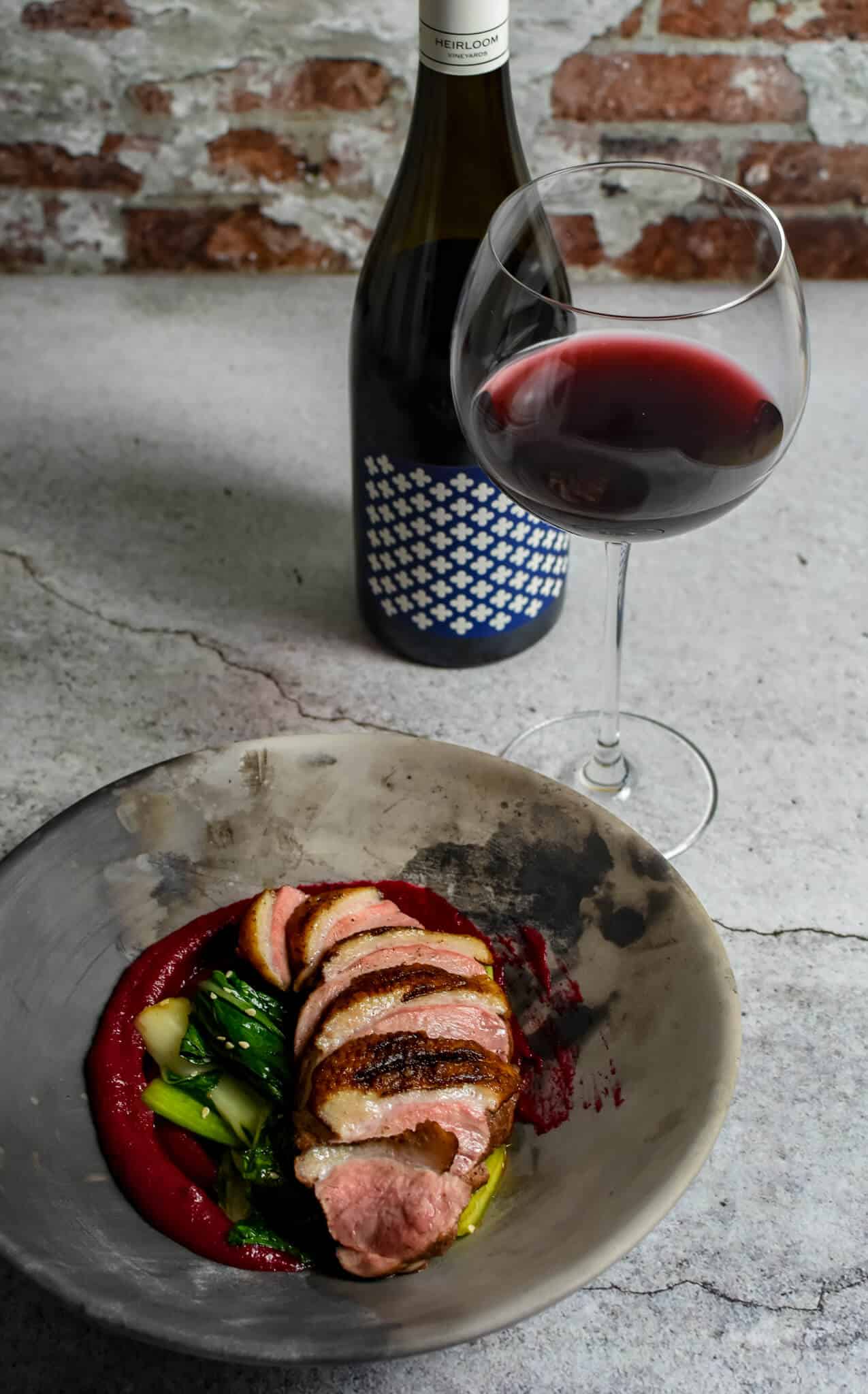 Duck breast, rhubarb ketchup & asian greens in a made of australia bowl on a table with heirloom pinot noir