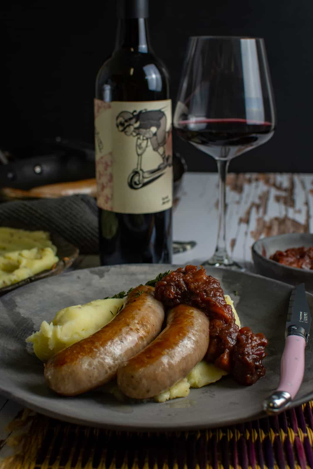 gourmet bangers and mash on a plate with a pink knife and red wine in the background