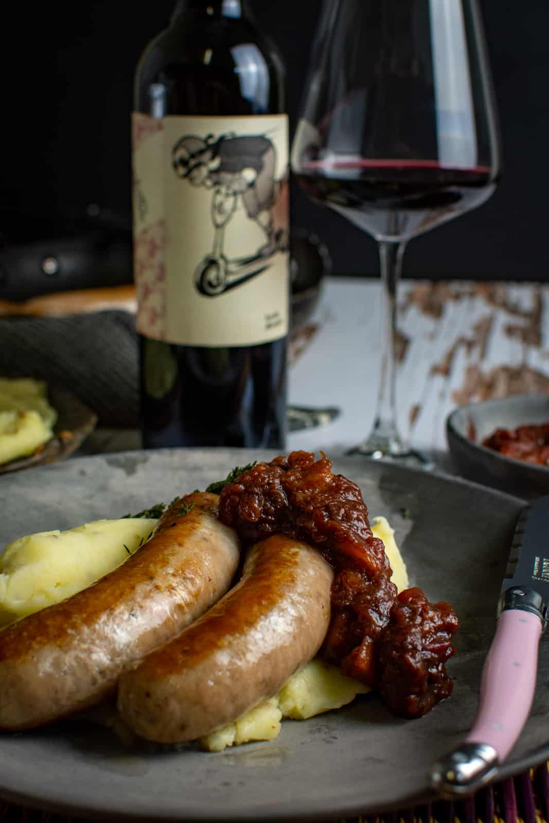 gourmet bangers and mash on a plate with a pink knife and red wine in the background