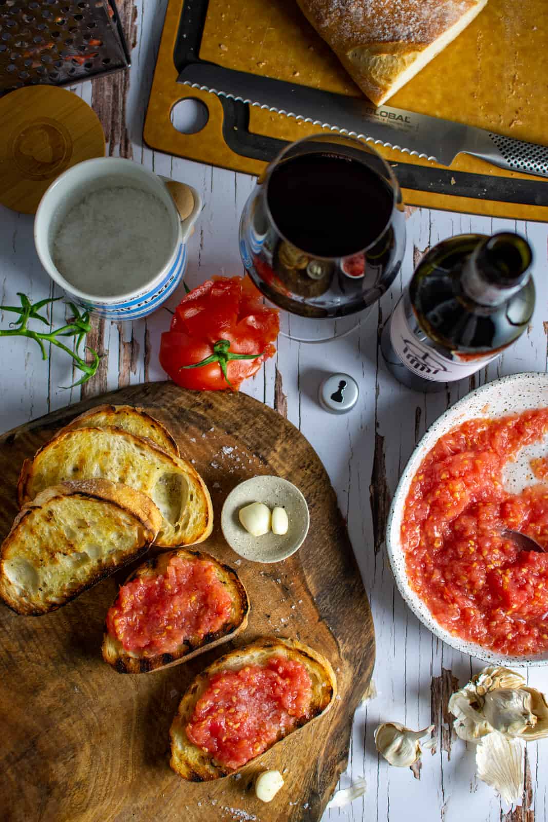 pan con tomate on a chopping board, garlic, tomatoes and wine on a table