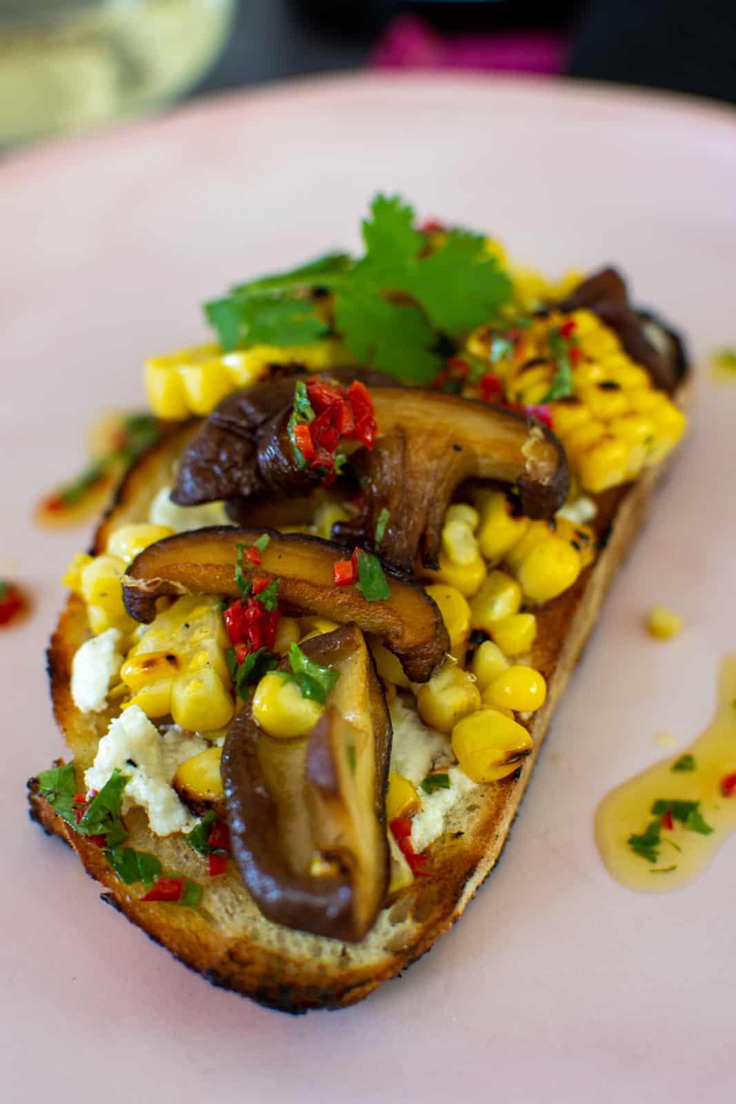 A slice of veggie toast topped with goat cheese, mushrooms, corn and coriander chilli oil on a plate