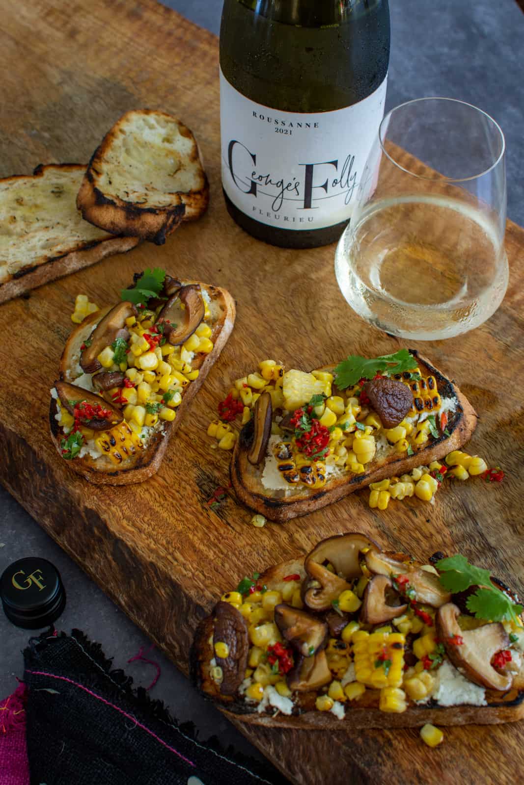 pieces of toast on wooden board topped with corn, mushroom & goat cheese. Bottle of white wine next to them 