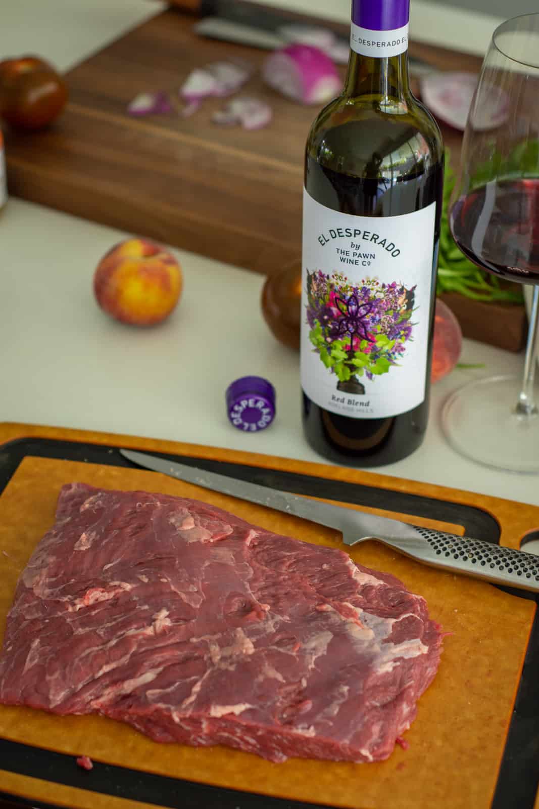 steak on chopping board, peaches and onions in background with a bottle of red wine beside