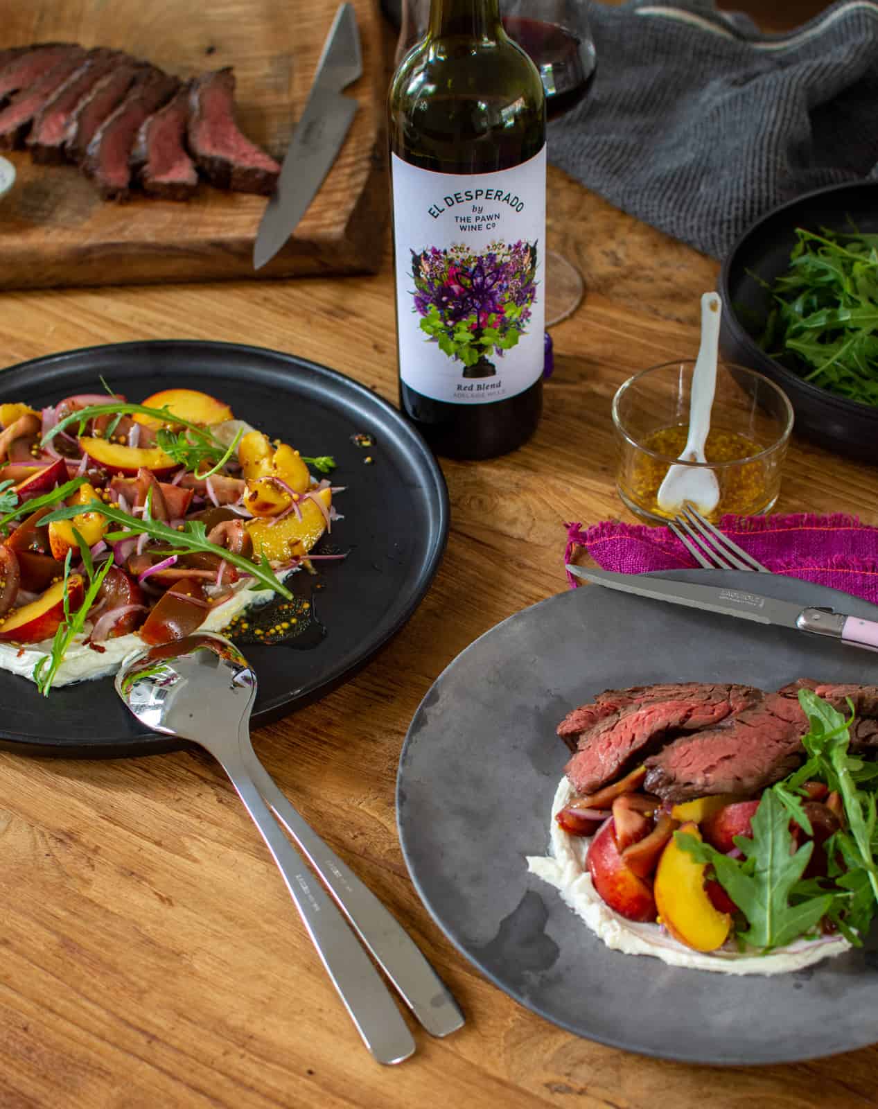 Plates of grilled bavette steak with peach, tomato salad on a table with bottle of red wine beside the plates