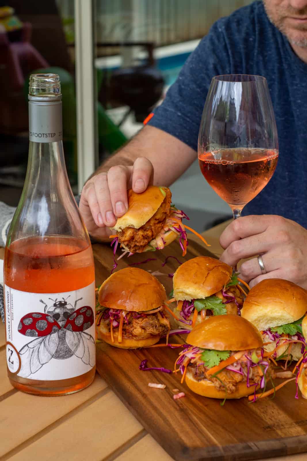 someone taking a fried chicken slider of a board whilst holding a glass of rose 