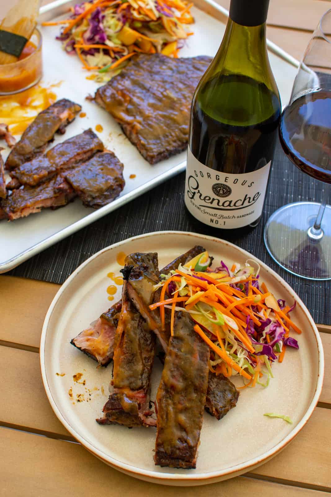 a table with a plate of smoked pork ribs, a tray of smoked pork ribs, slaw and red wine 