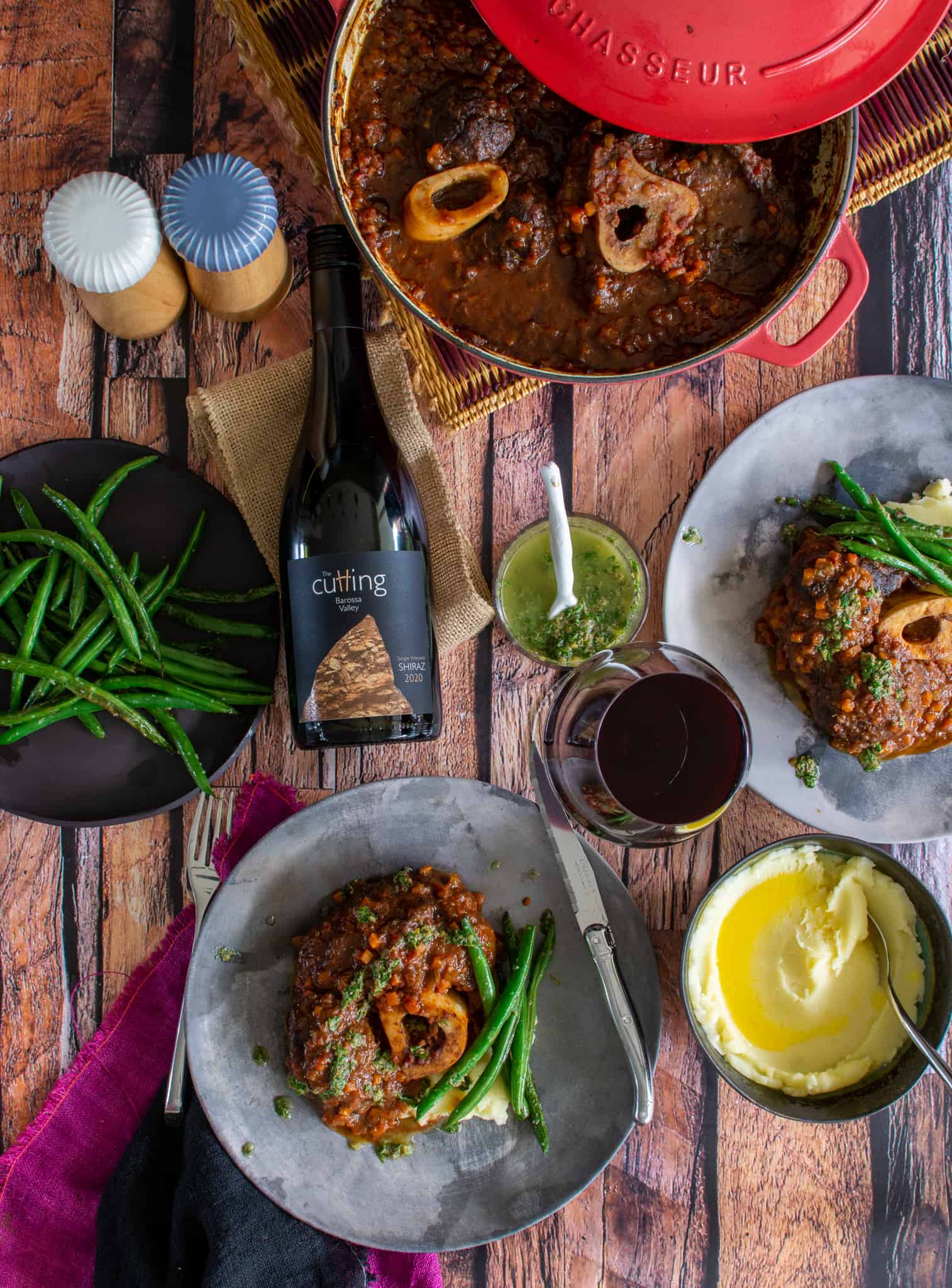 Beef ossobuco on a table with red wine, wine glasses and other food on plates and in bowls