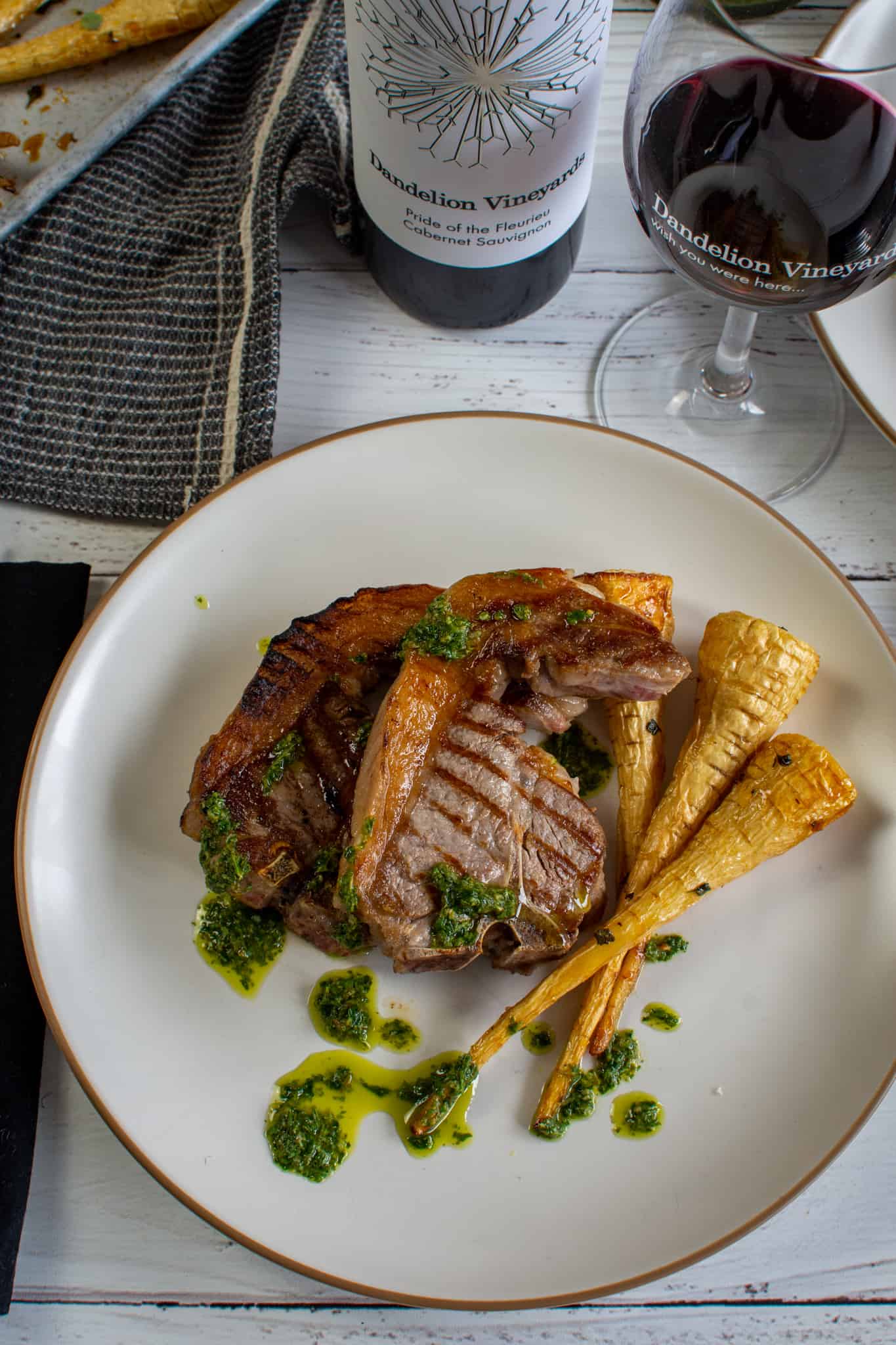 grilled lamb loin chops, salsa verde nd roasted parsnips on a plate with a bottle of wine behind the plate