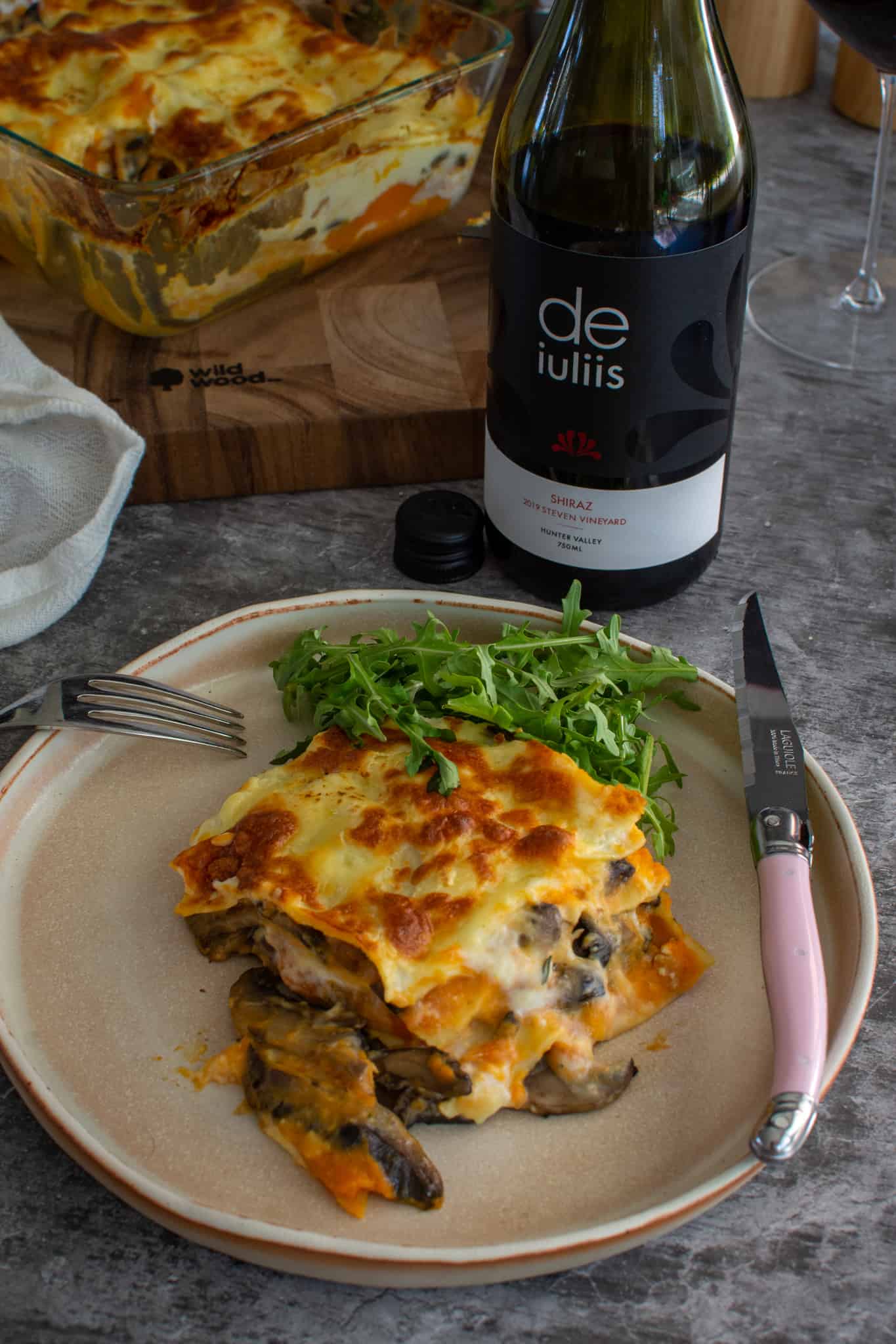 Vegetable lasagne on a plate with a small bit of rocket salad too.  De iuliis wine beside it with lasagne dish in background 