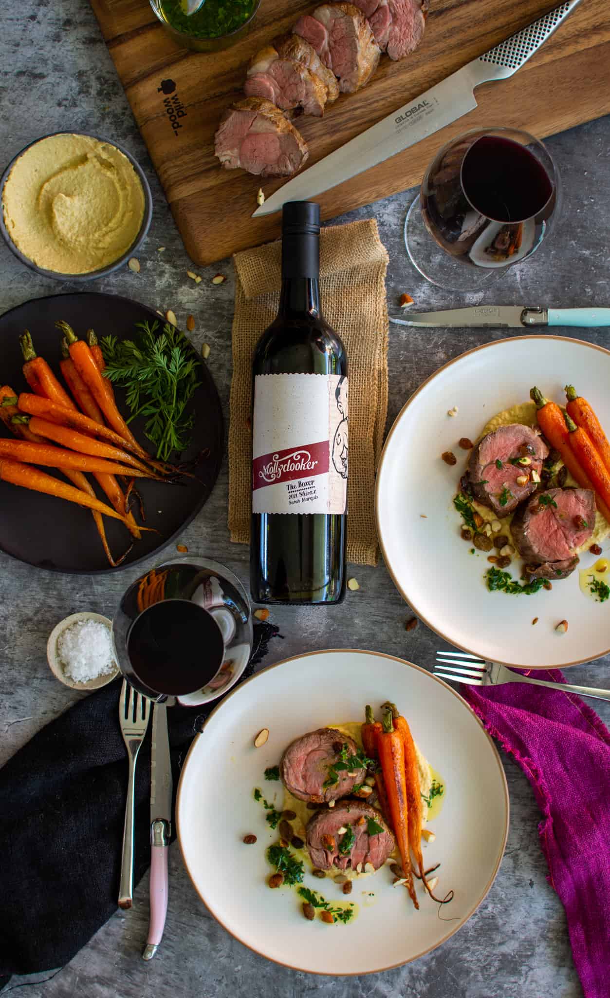 roasted bonless lamb shoulder with hummus and carrots on a plate. Mollydoooker wine, cutlery and plate of carrots also on the table 